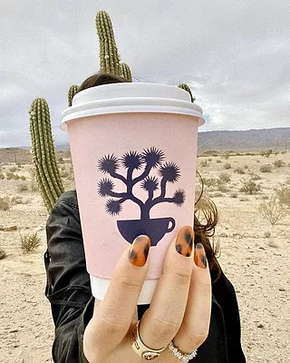 Woman holding a pink paper cup of coffee over her face with a cactus behind her.