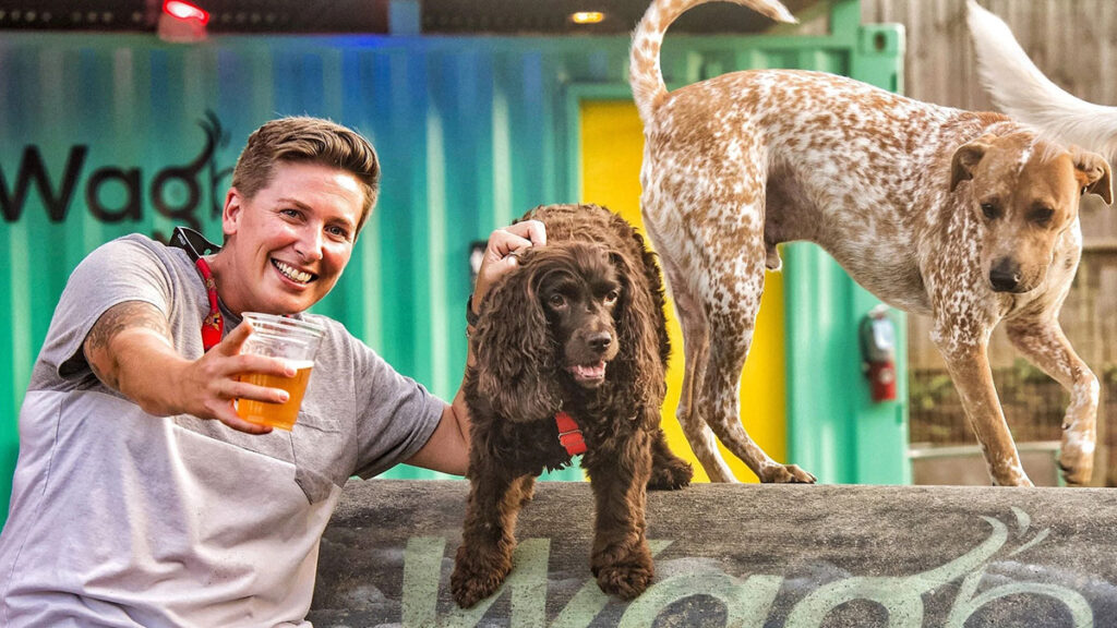 smiling woman holding a beer with two dogs next to her.
