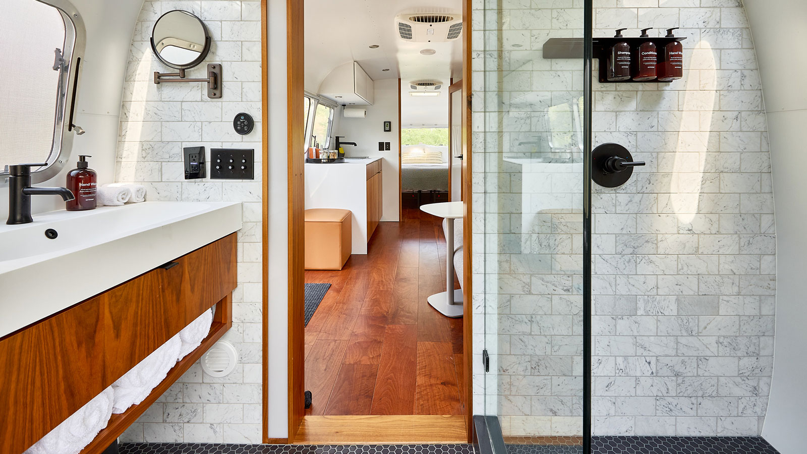 Inside view of an Airstream showing kitchen and bathroom with rich wood floor and white marble tiles