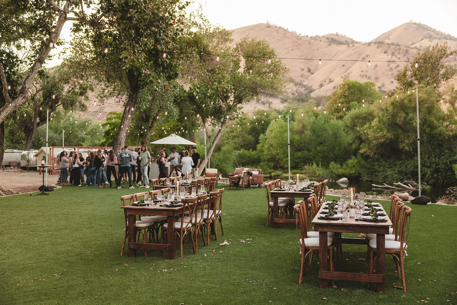 Group meeting in an outdoor area with tables set up with candles. Trees and golden hills in the background and string lights overhead at AutoCamp Sequoia. Known as Ponderosa Lawn.