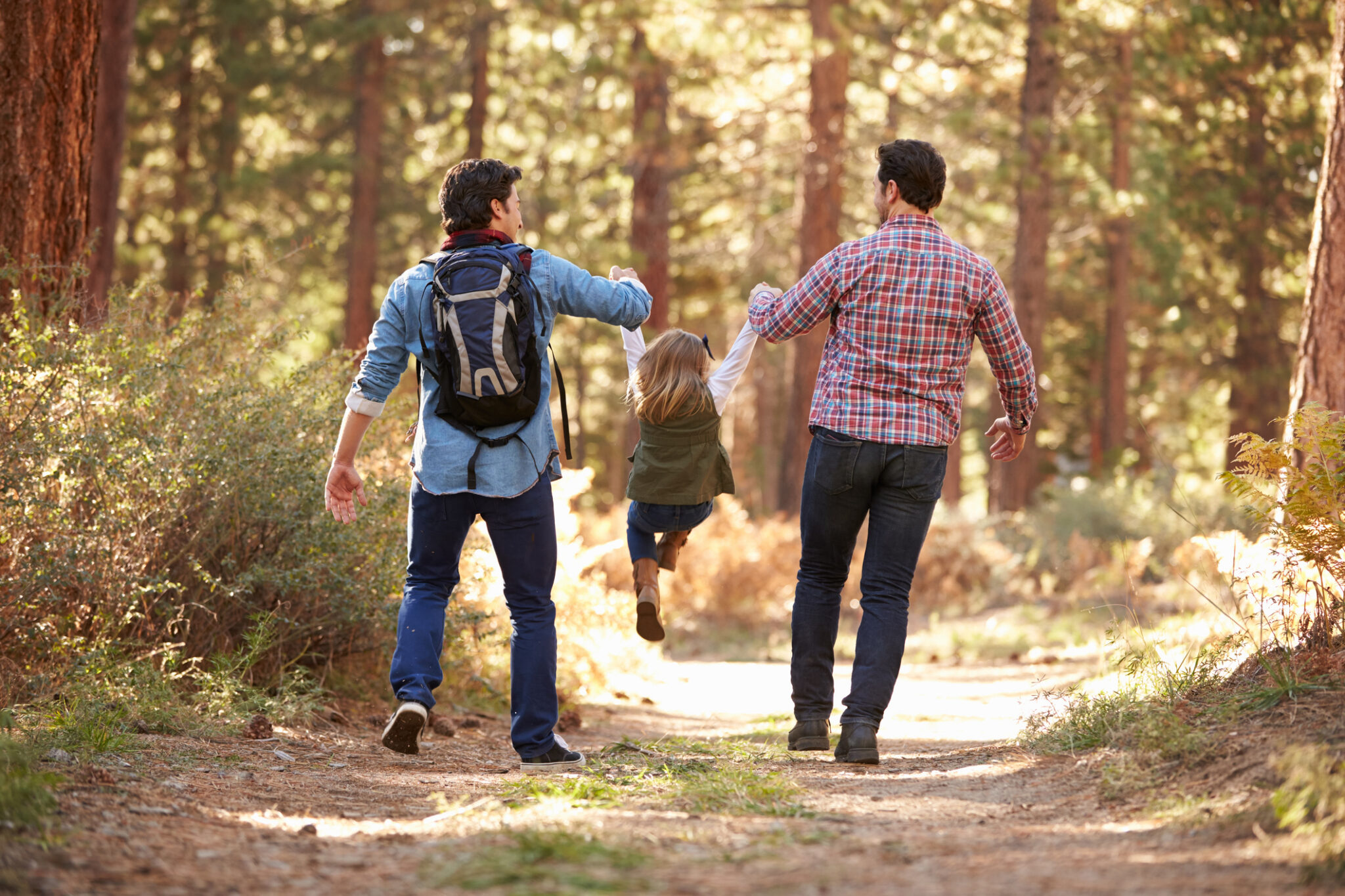 Two dads hiking lifting their young daughter between them on the path